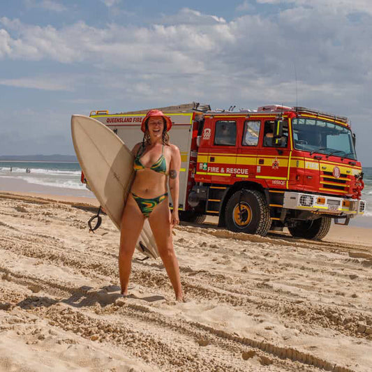 Summer from SunClub at Rainbow Beach with her surfboard and a fire truck, wearing the SunClub x Plow surf hat