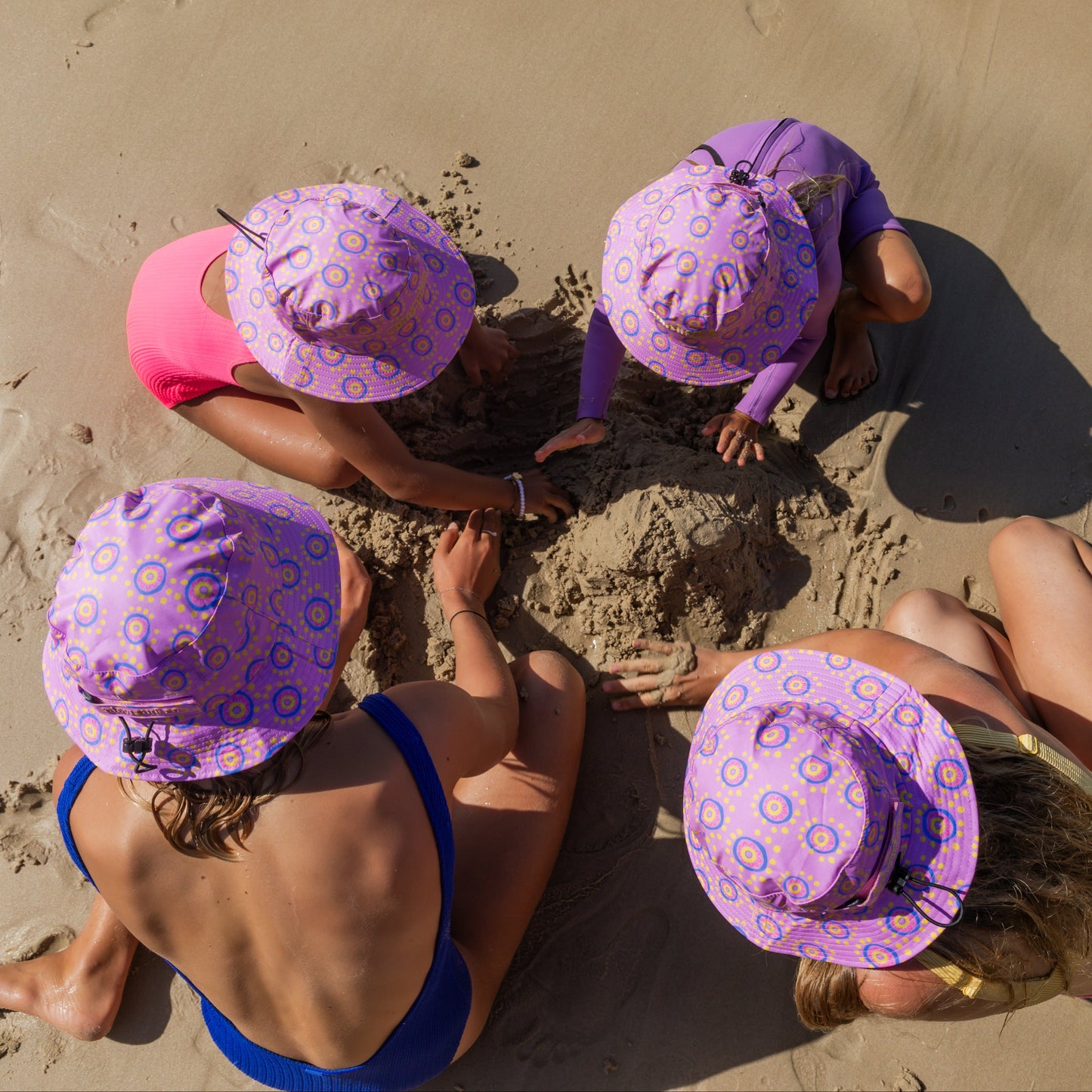 Four people sitting on a sandy beach wearing colorful surf hats by plow surf co. x the minishakas leihani zoric