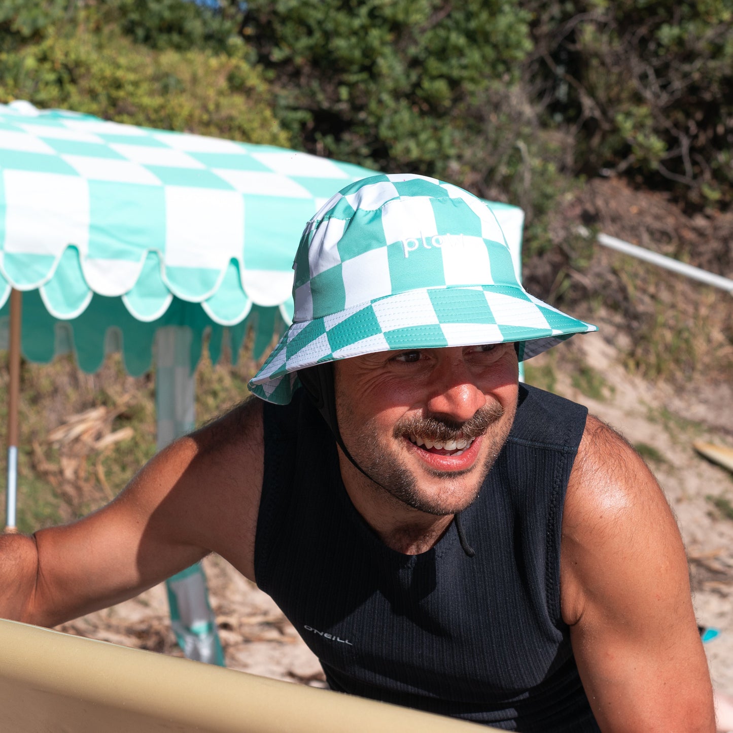 Man with a mint coloured checkered surf hat on his head holding a surfboard outdoors