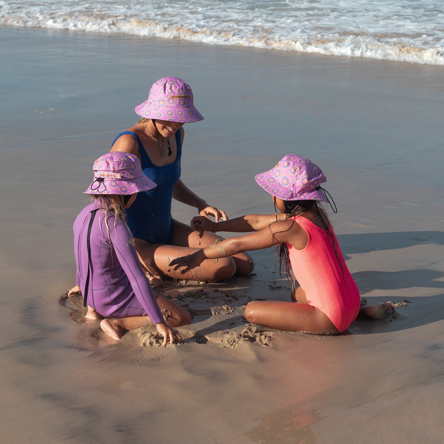 Three children in swimsuits and plow surf hats playing in the sand on a beach.