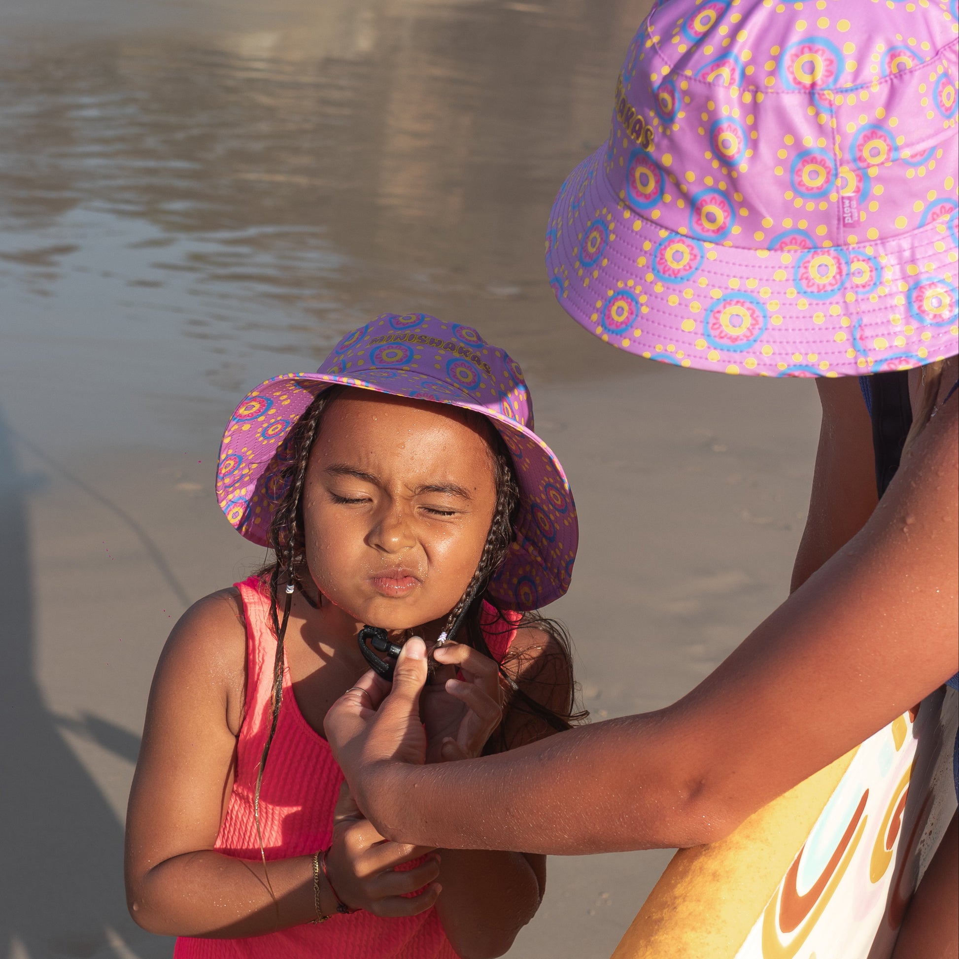 Two children wearing purple hats on a beach with a surfboard.