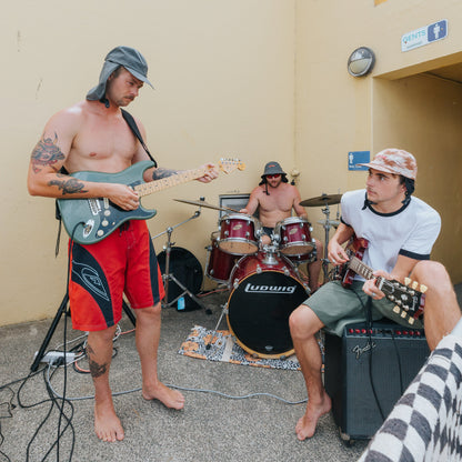 Three men playing music in a casual setting with a beige wall. Wearing plow surf hats in charcoal colour & camo print NEVER forget protection
