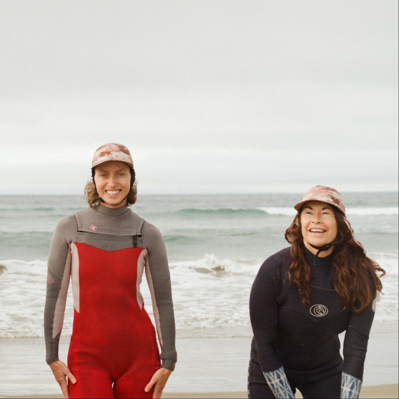 2 women in wetsuits standing on a beach with ocean in the background wearing plow surf hats in Camo NEVER forget protection style surf caps