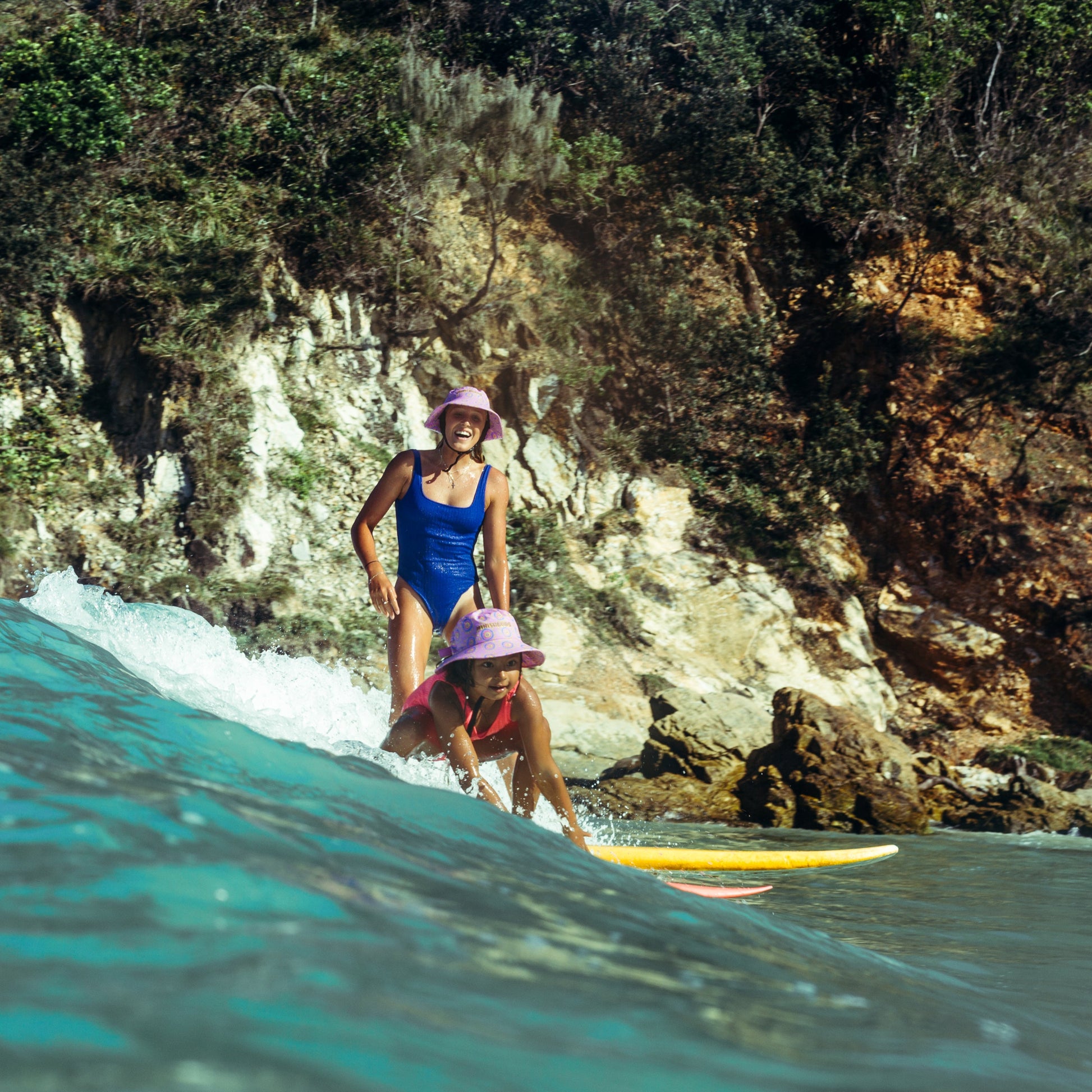 Two people on surfboards in the ocean with a rocky cliff and vegetation in the background wearing plow surf hats designed by leihani zoric the minishakas