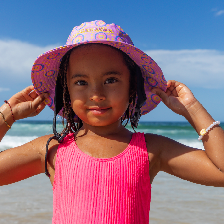 Child in a pink colorful surf hat by plow surf co. designed by Leihani Zoric the minishakas to support her aboriginal art and surfing goals